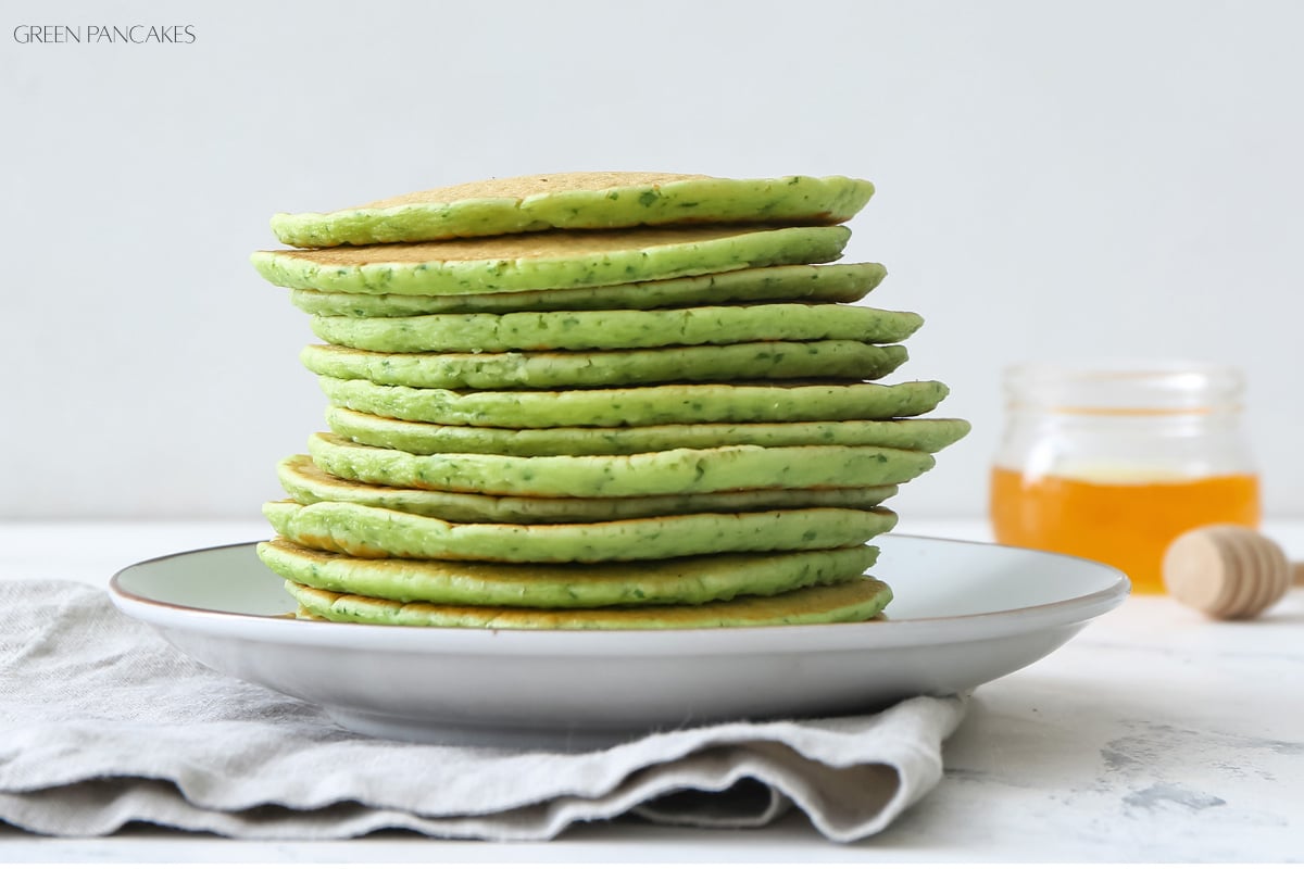 A stack of green pancakes sits on a white plate with a jar of honey and a wooden honey dipper in the background.