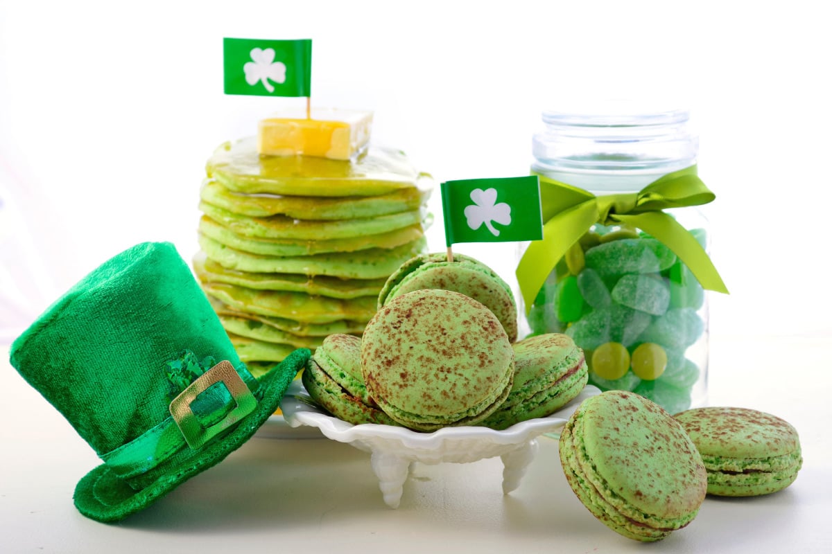 A stack of green pancakes with a shamrock flag, green macarons, a jar of green candy, and a green leprechaun hat arranged on a table.
