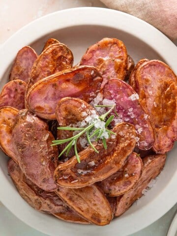 A bowl of roasted purple potatoes garnished with coarse salt and a sprig of rosemary.