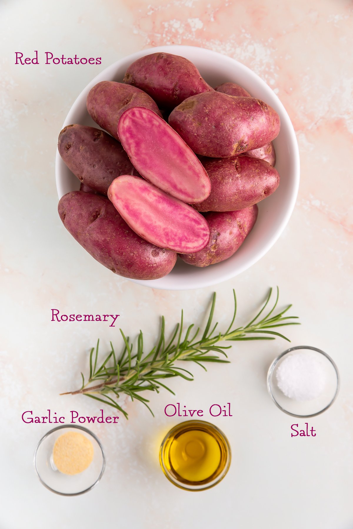 A bowl of red potatoes, a sprig of rosemary, a small glass bowl of garlic powder, a small glass bowl of olive oil, and a small bowl of salt on a light surface.