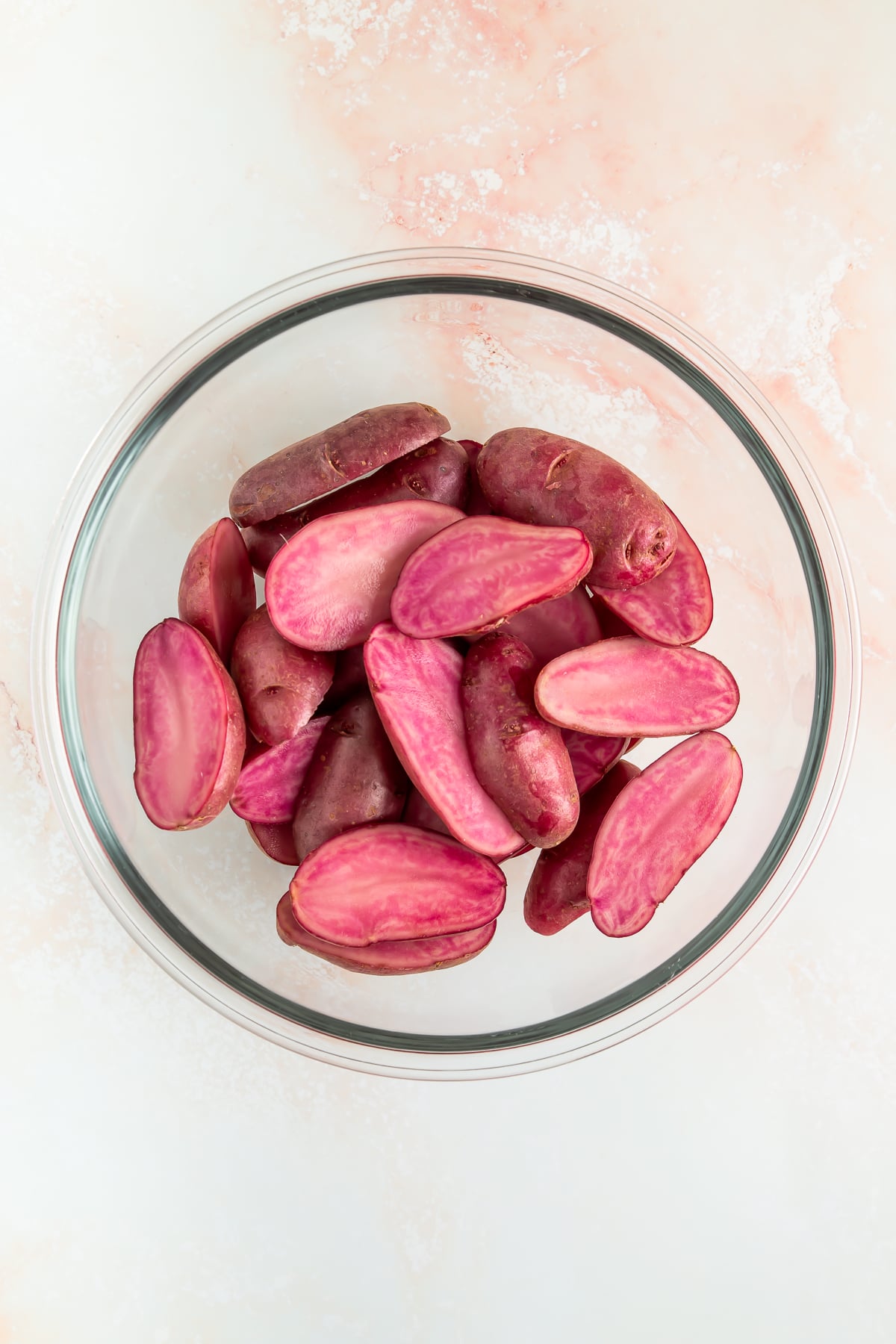 A glass bowl filled with halved purple potatoes on a light pink and white surface.