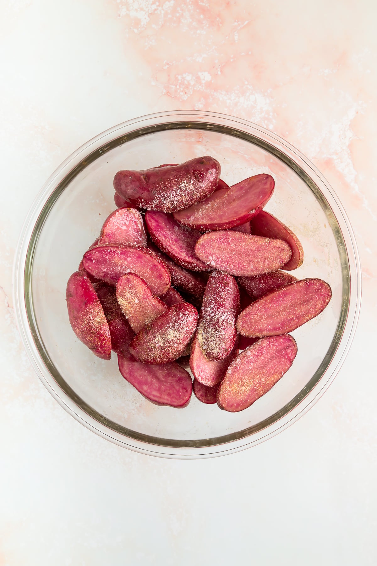 A glass bowl filled with sliced purple potatoes, sprinkled with seasoning, on a light-colored surface.