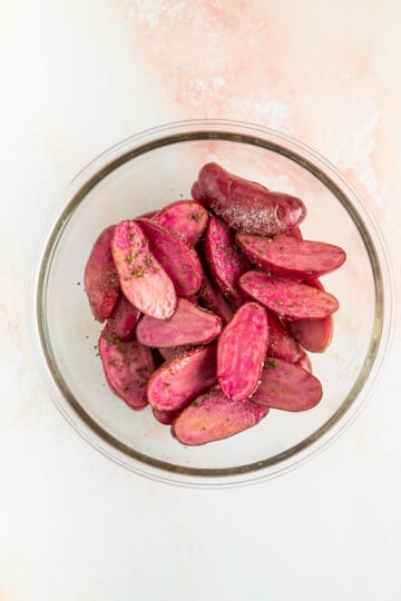 A glass bowl filled with halved purple potatoes seasoned with herbs and spices on a light-colored surface.