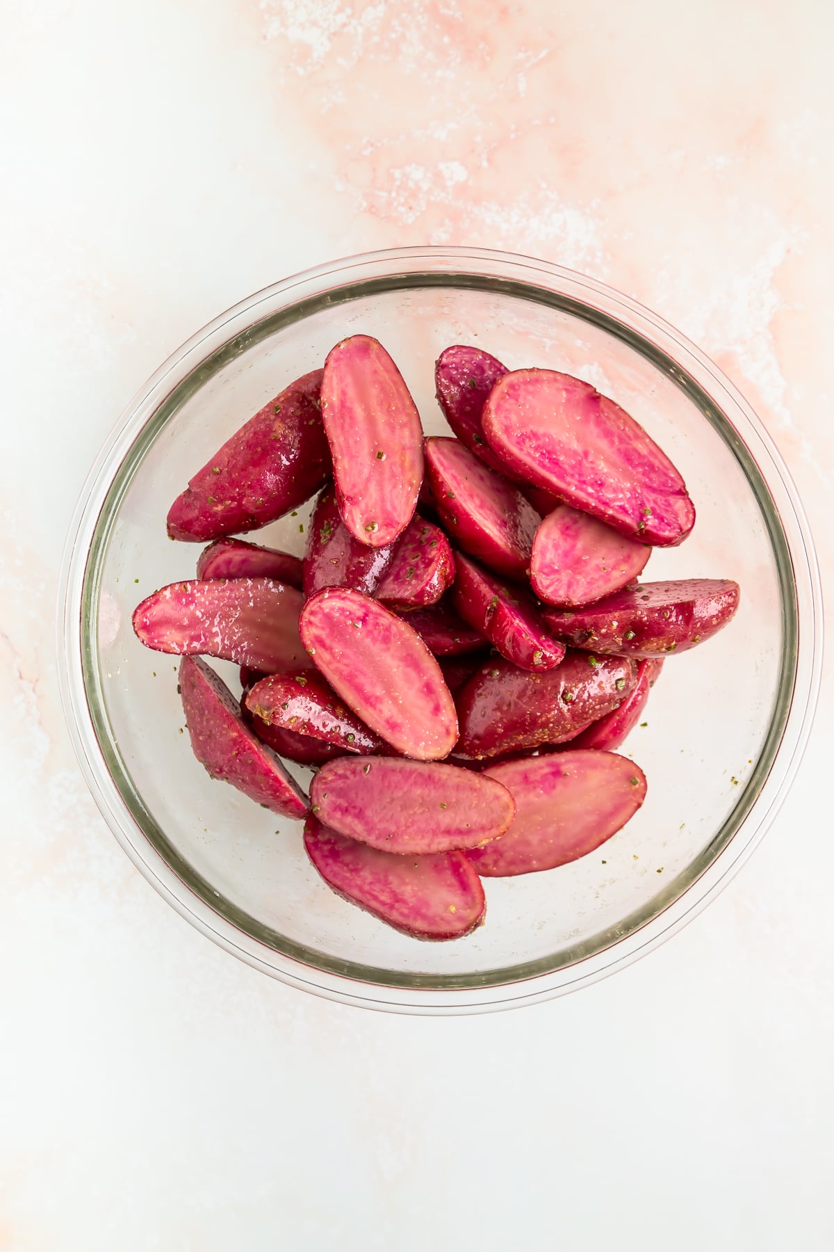 A glass bowl filled with halved red potatoes seasoned with pepper on a light pink and white surface.