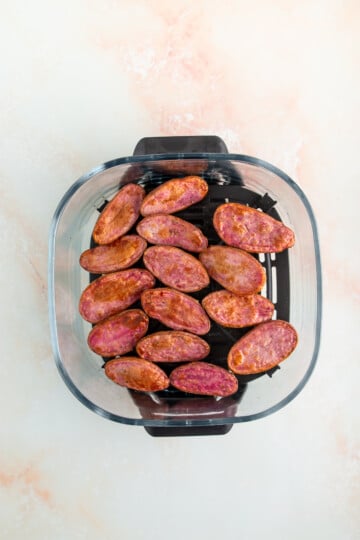 Halved seasoned purple potatoes arranged in a single layer inside the basket of an air fryer on a light-colored surface.
