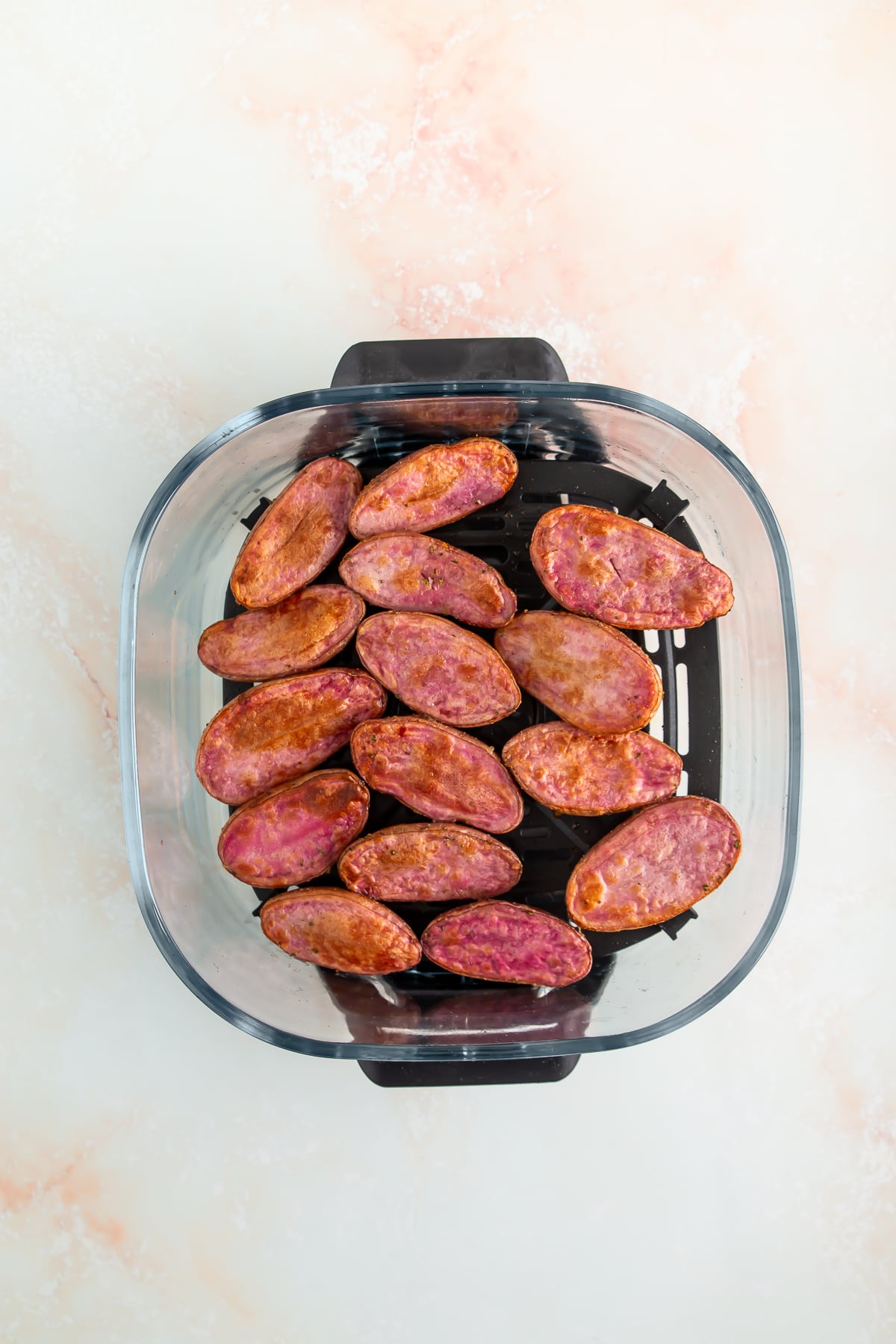 Halved seasoned purple potatoes arranged in a single layer inside the basket of an air fryer on a light-colored surface.