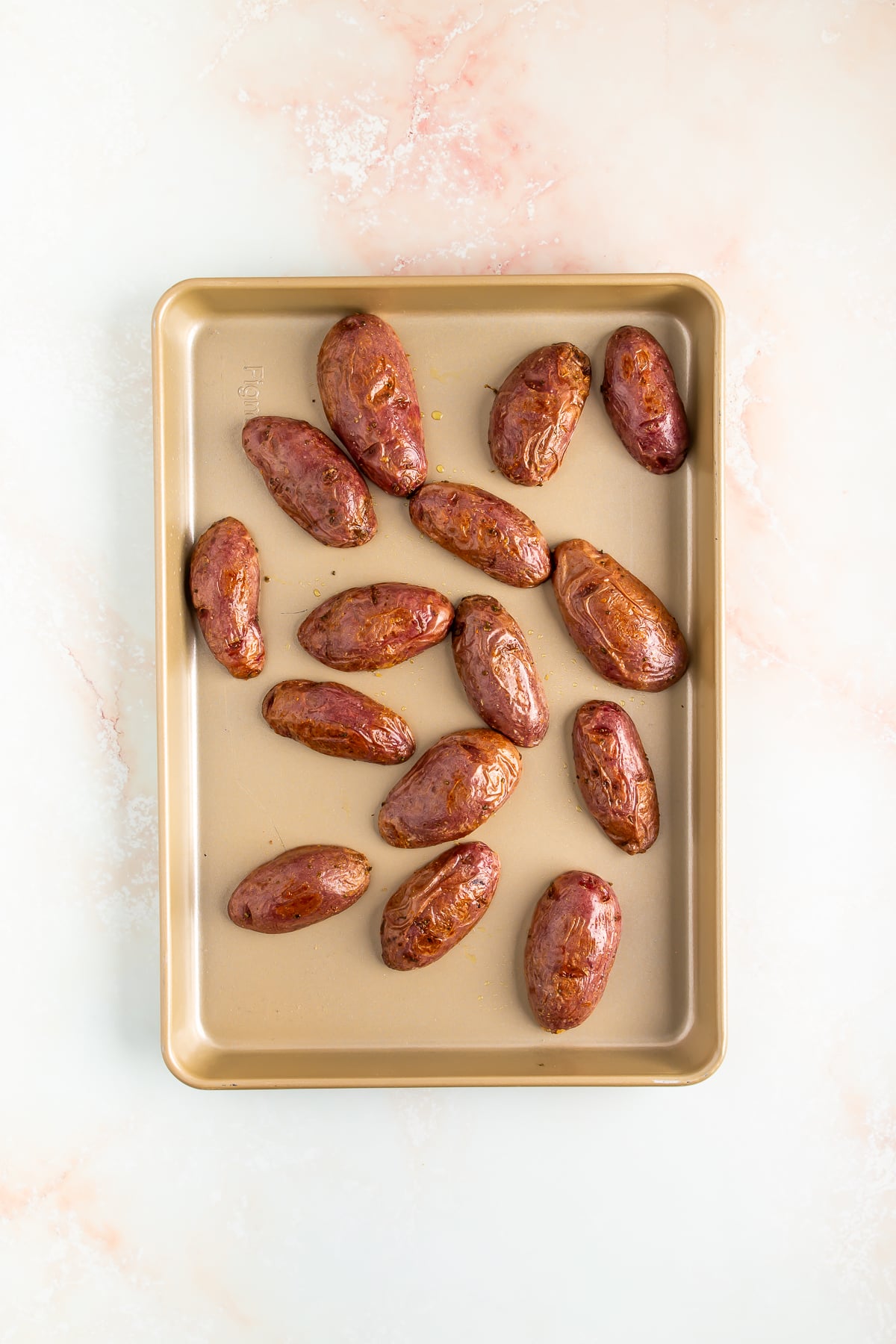 A gold baking sheet with fifteen whole, unpeeled red potatoes, spaced apart, on a light marbled surface.