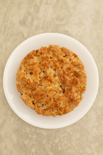 A round, baked oatmeal cookie with a golden-brown top sits on a white plate against a light, textured background.