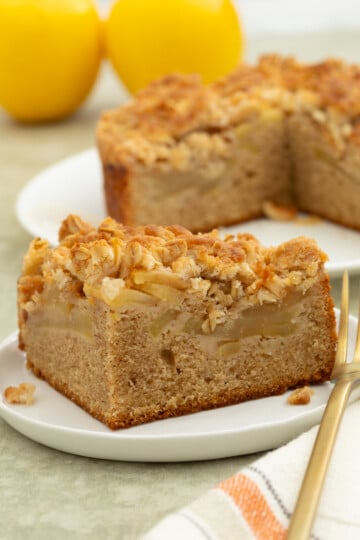 A slice of apple crumb cake with a crumb topping sits on a white plate, with the remaining cake and two yellow apples in the background.