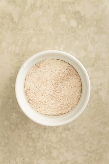 A white bowl filled with light beige flour sits on a textured, light-colored surface.