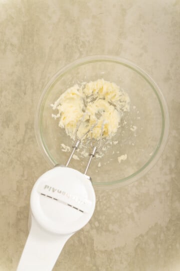 A hand mixer creaming butter in a clear glass bowl on a light-colored countertop.
