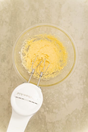 A hand mixer is blending yellow batter in a glass bowl on a light-colored countertop.