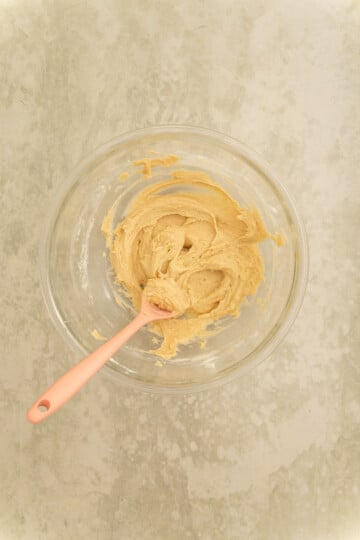 A glass bowl with light brown cookie dough and a pink spatula rests on a light-colored textured surface.