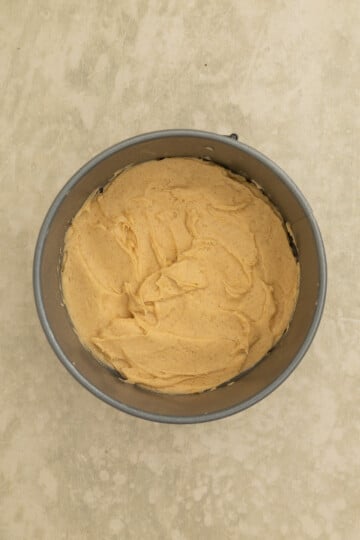 A round baking pan filled with smooth, light brown cake batter, placed on a beige surface.