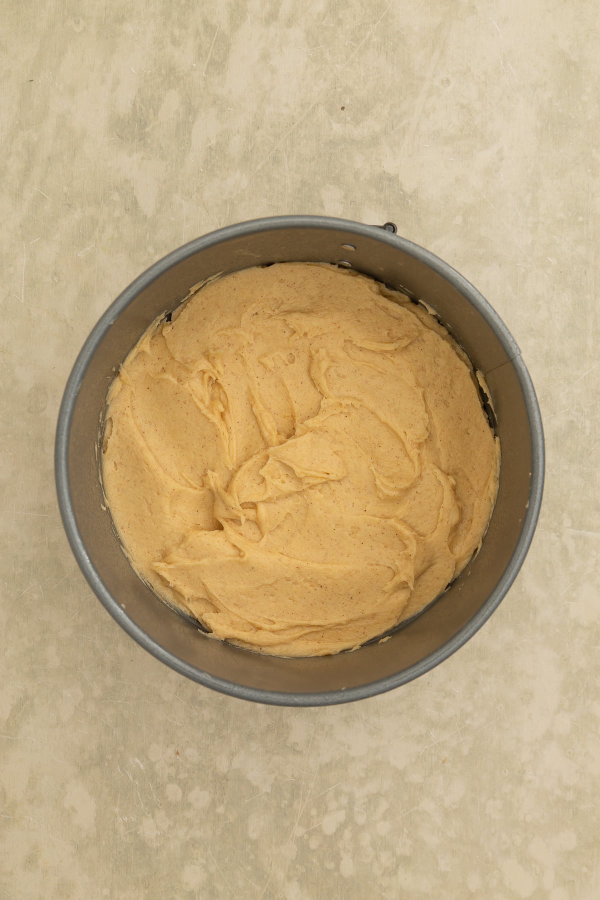 A round baking pan filled with smooth, light brown cake batter, placed on a beige surface.