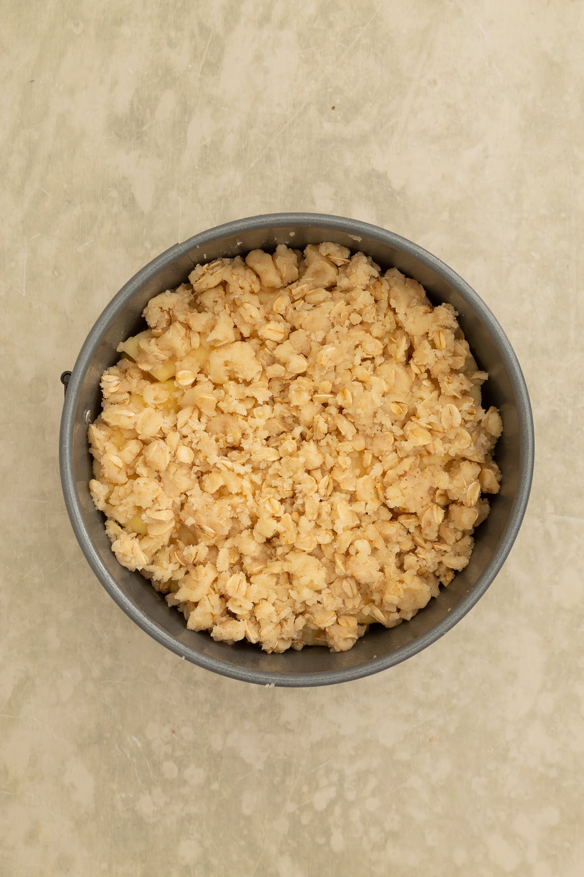 Round baking pan filled with an unbaked crumbly mixture, likely a crumble or streusel topping, on a light-colored textured surface.