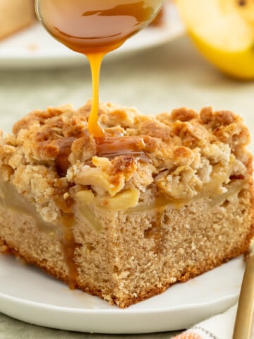 A slice of apple crumb cake on a plate is topped with a drizzle of caramel sauce. A fork and a napkin are placed beside the plate.