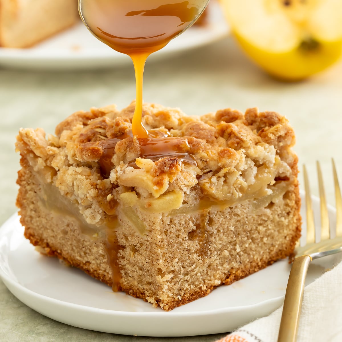 A slice of apple crumb cake on a plate is topped with a drizzle of caramel sauce. A fork and a napkin are placed beside the plate.