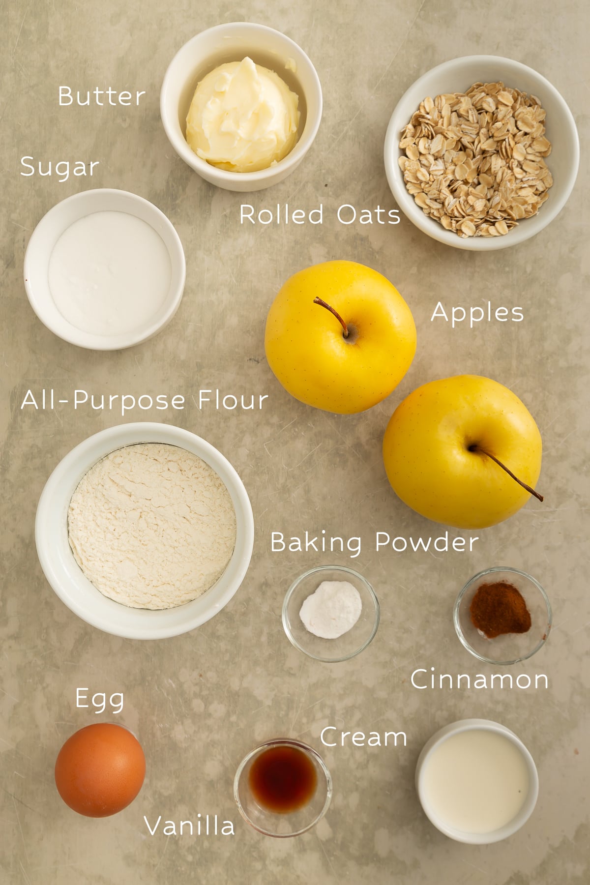 A top view of ingredients for baking, including apples, butter, rolled oats, sugar, flour, baking powder, cinnamon, egg, vanilla, and cream, each labeled.