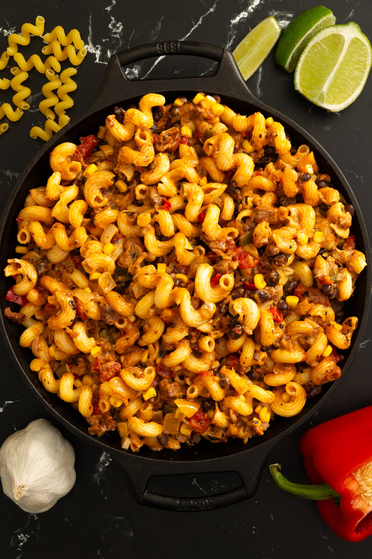 A skillet filled with cooked cavatappi pasta, ground meat, corn, and tomatoes, surrounded by uncooked pasta, garlic, red bell pepper, and lime wedges on a black marble surface.