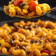 A close-up of a spoonful of pasta with black beans, corn, and red bell peppers, held above a skillet filled with the same pasta dish.