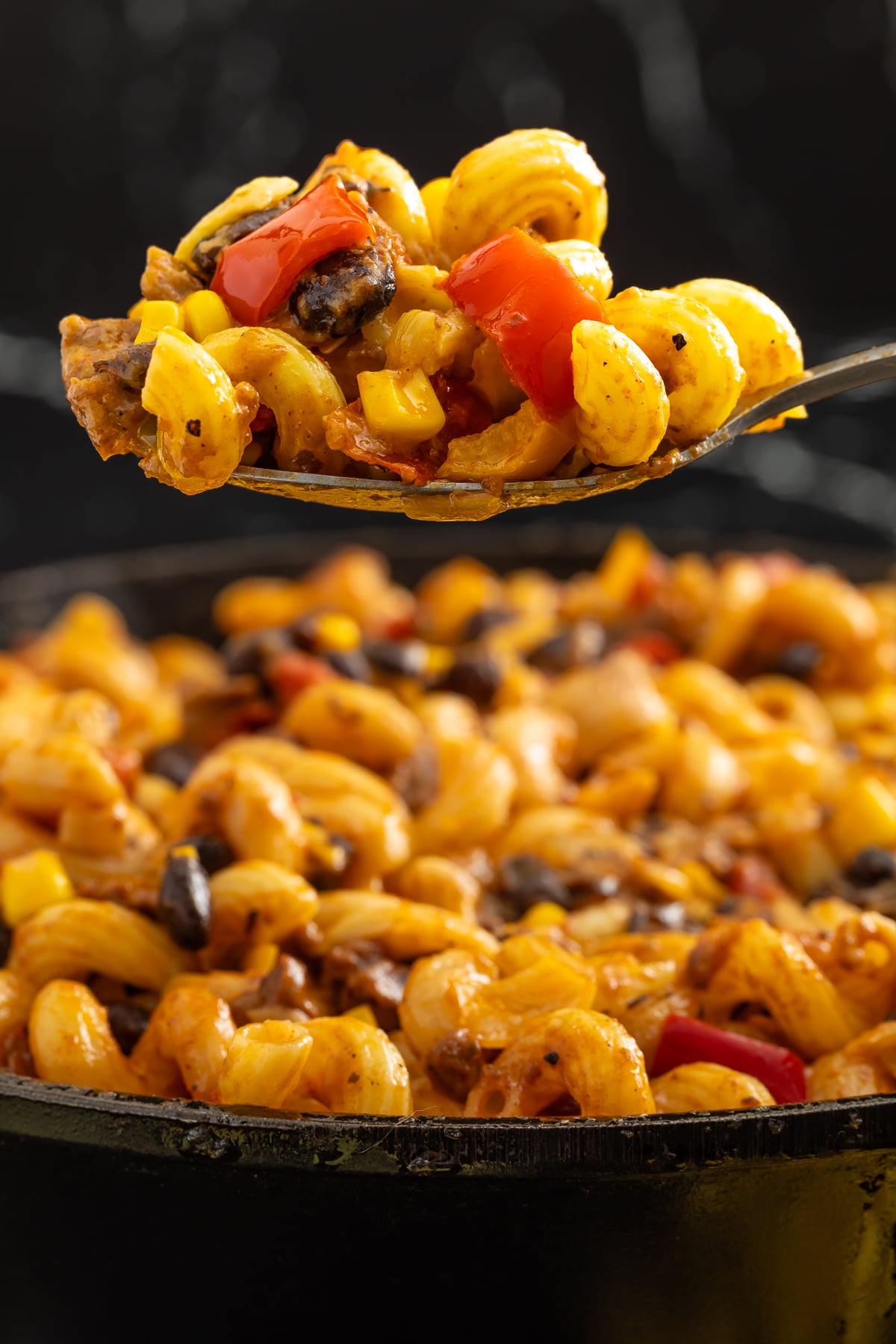 A close-up of a spoonful of pasta with black beans, corn, and red bell peppers, held above a skillet filled with the same pasta dish.