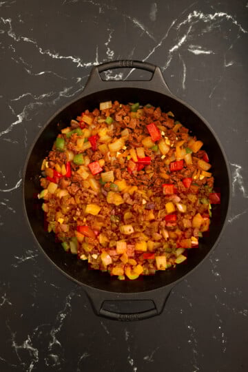 A black cast iron skillet filled with a cooked mixture of diced potatoes, bell peppers, onions, and ground meat on a dark marble surface.
