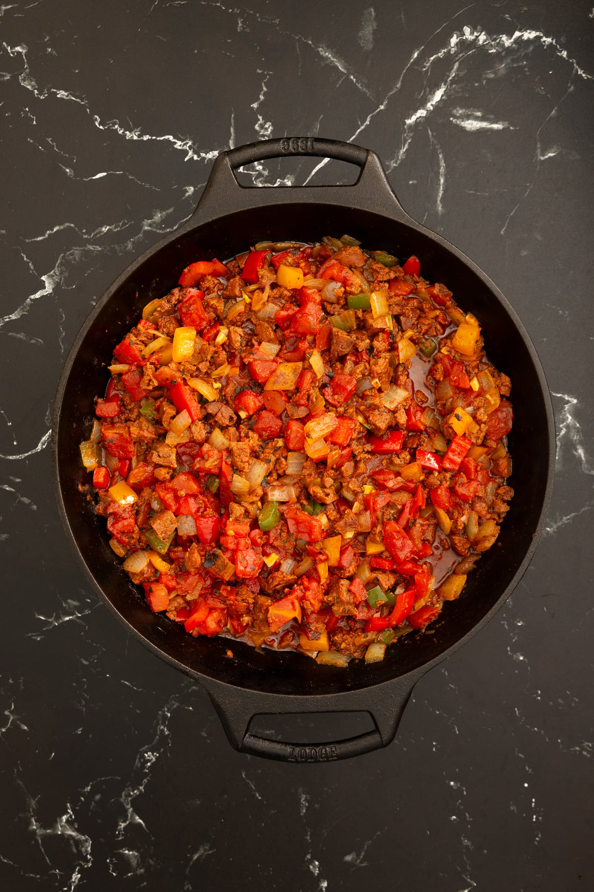 A black pot filled with a cooked mixture of chopped tomatoes, peppers, onions, and ground meat sits on a dark marble surface.
