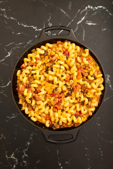 A black pan filled with cooked cavatappi pasta mixed with ground meat, tomatoes, onions, and seasonings on a black marble surface.