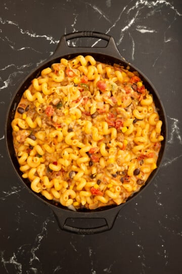 A cast iron skillet filled with cooked cavatappi pasta mixed with cheese, black beans, tomatoes, and sauce sits on a black marble countertop.