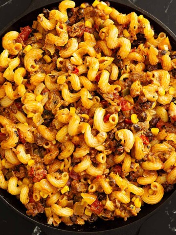 A skillet filled with cooked cavatappi pasta mixed with ground beef, corn, tomatoes, and spices on a black marble surface.
