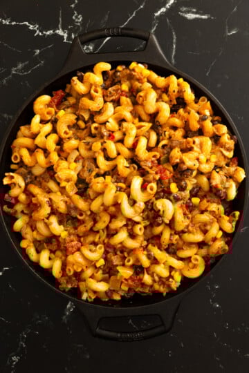 A skillet filled with cooked cavatappi pasta mixed with ground beef, corn, and diced tomatoes on a black marble surface.
