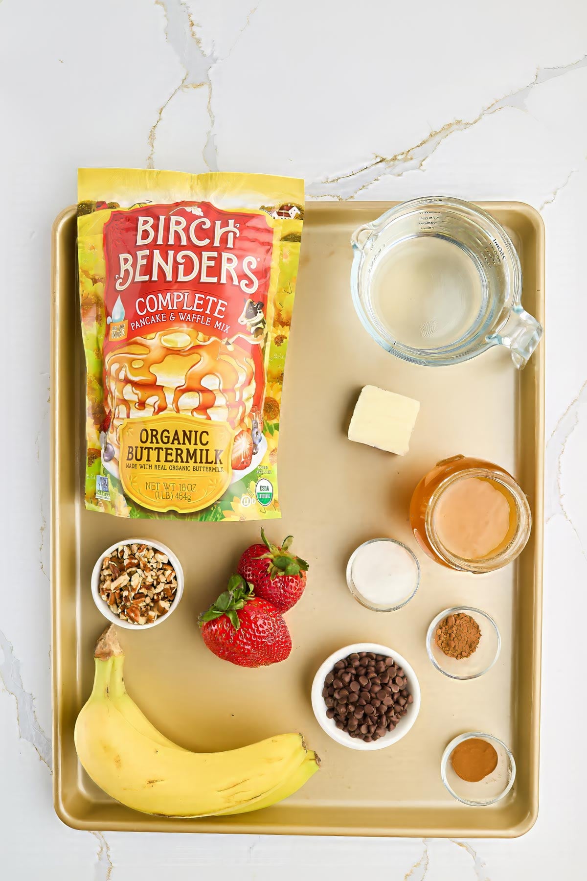 A tray with Birch Benders pancake mix, water in a measuring cup, butter, honey, strawberries, a banana, chopped nuts, chocolate chips, salt, cinnamon, and baking powder in small bowls.