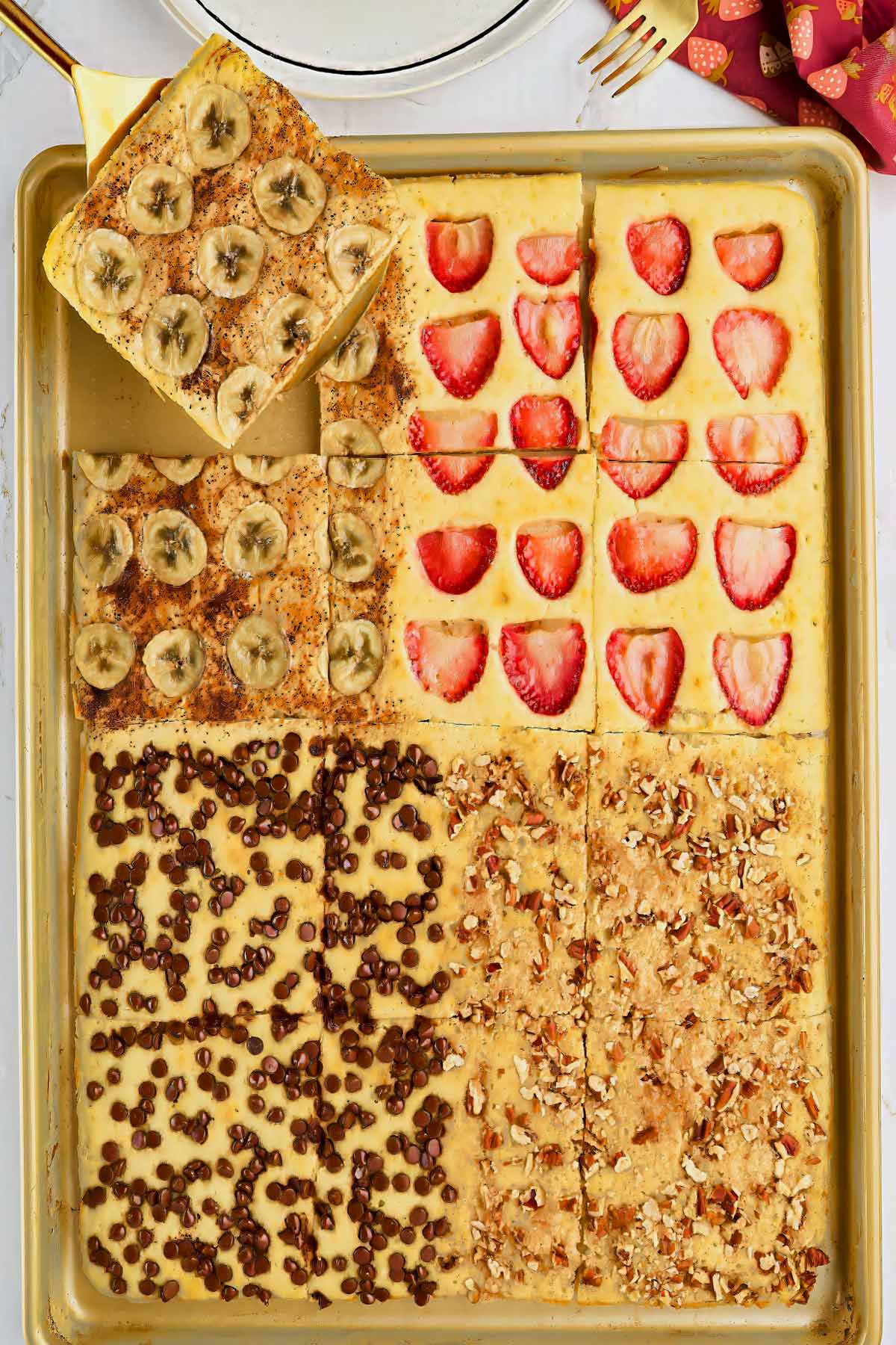 A baking sheet with pancake squares, each topped differently: banana slices, strawberries, chocolate chips, and chopped nuts. One banana slice square is being lifted with a spatula.