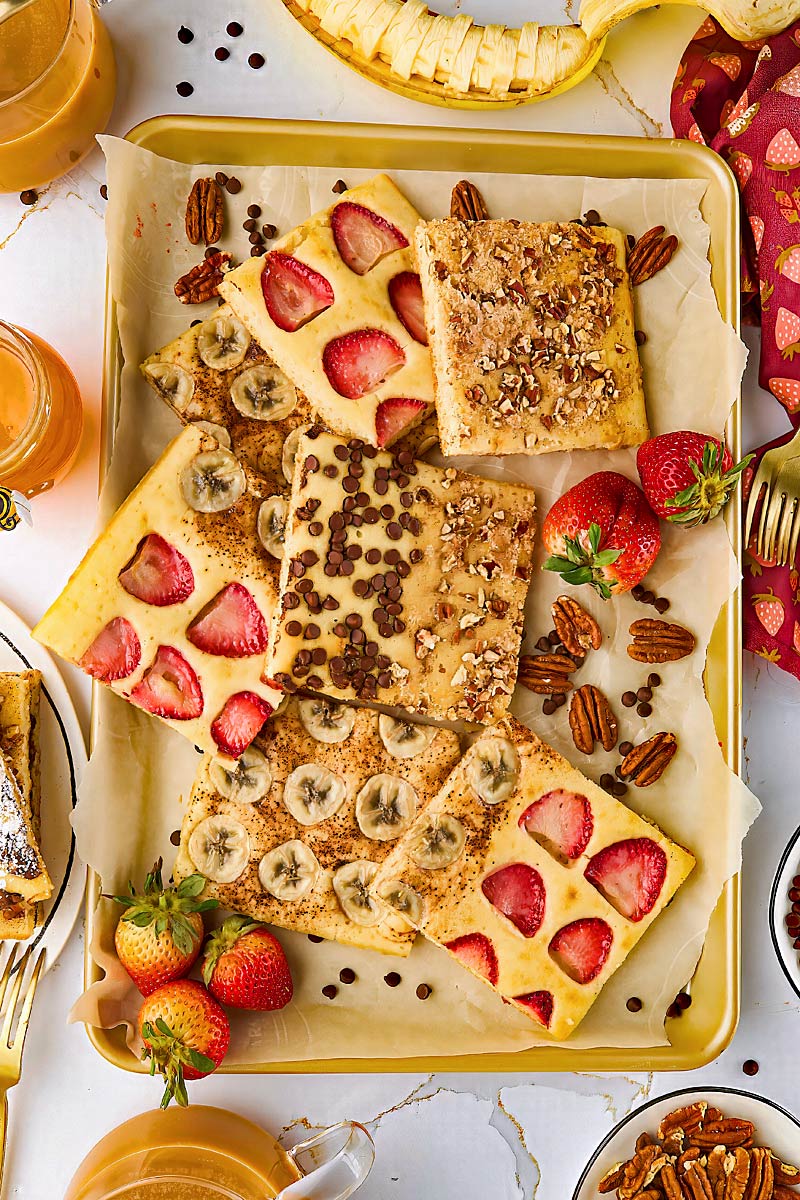 A tray with six baked pancake squares topped with sliced strawberries, bananas, chocolate chips, and chopped nuts, surrounded by fresh strawberries and pecans.