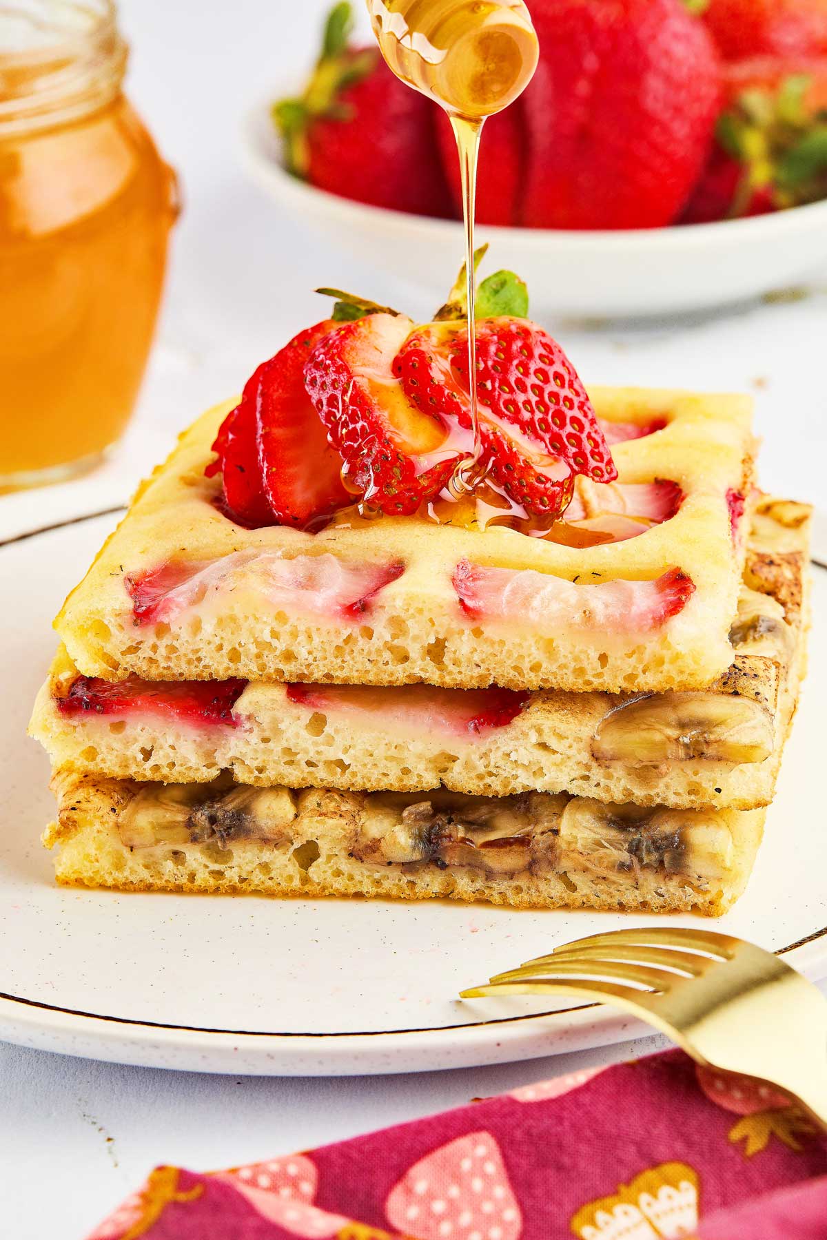 A stack of strawberry and banana sheet pancakes topped with sliced strawberries, with honey being drizzled on top. A jar of honey and a bowl of strawberries are in the background.