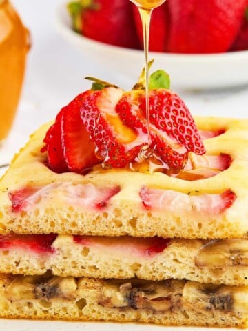 Three stacked sheet pan pancakes from mix with strawberry and banana slices, topped with fresh strawberries and honey being drizzled on top. A bowl of strawberries and a jar of honey sit in the background.