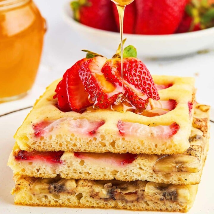 Three stacked sheet pan pancakes from mix with strawberry and banana slices, topped with fresh strawberries and honey being drizzled on top. A bowl of strawberries and a jar of honey sit in the background.