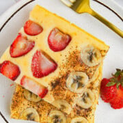 Two square pieces of baked pancake, one topped with sliced strawberries and the other with sliced bananas, are plated with a whole strawberry and a gold fork on a white plate.
