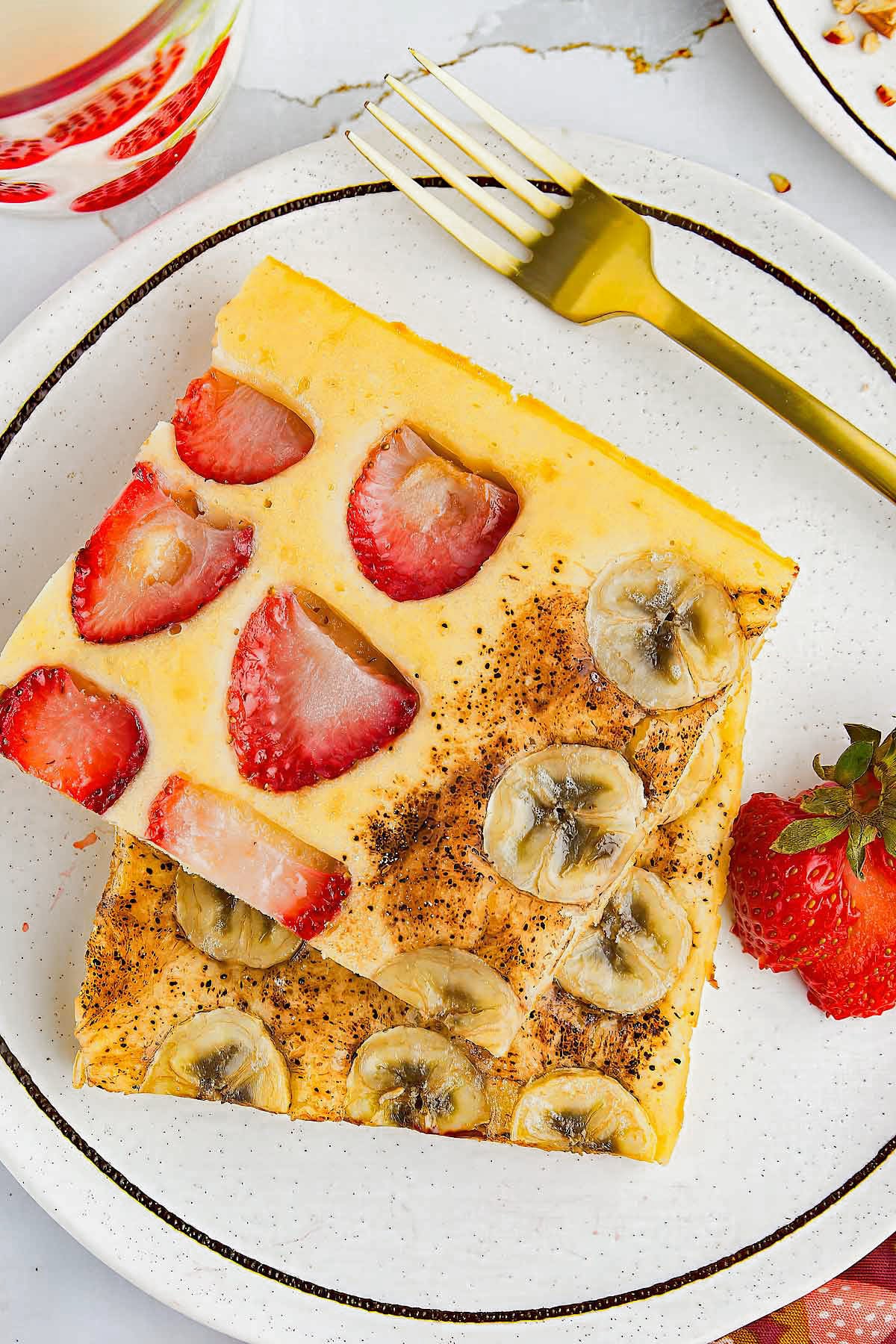 Two square pieces of baked pancake, one topped with sliced strawberries and the other with sliced bananas, are plated with a whole strawberry and a gold fork on a white plate.