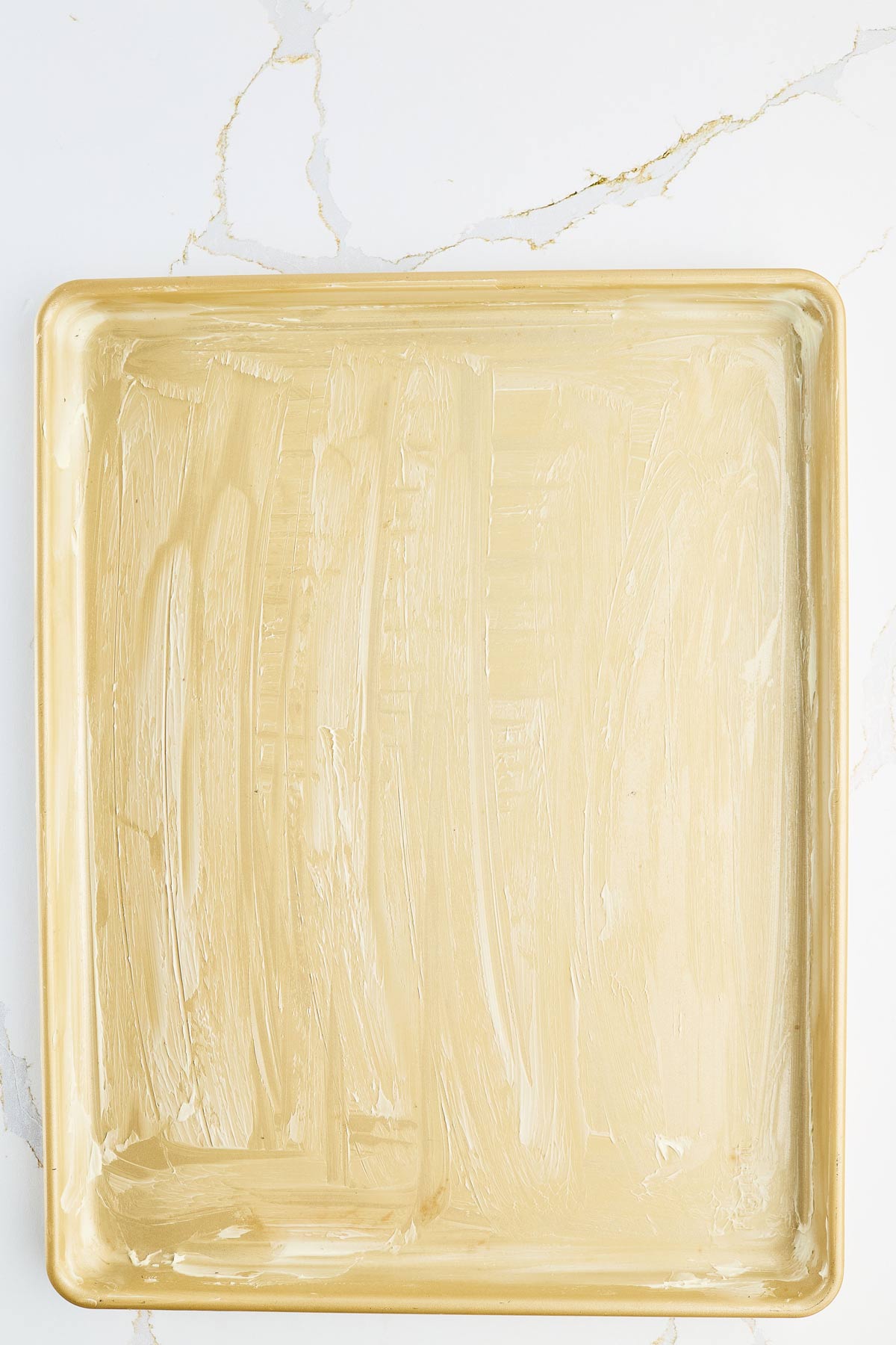 A gold-colored rectangular baking sheet is coated with a thin, even layer of butter on a white marble surface.