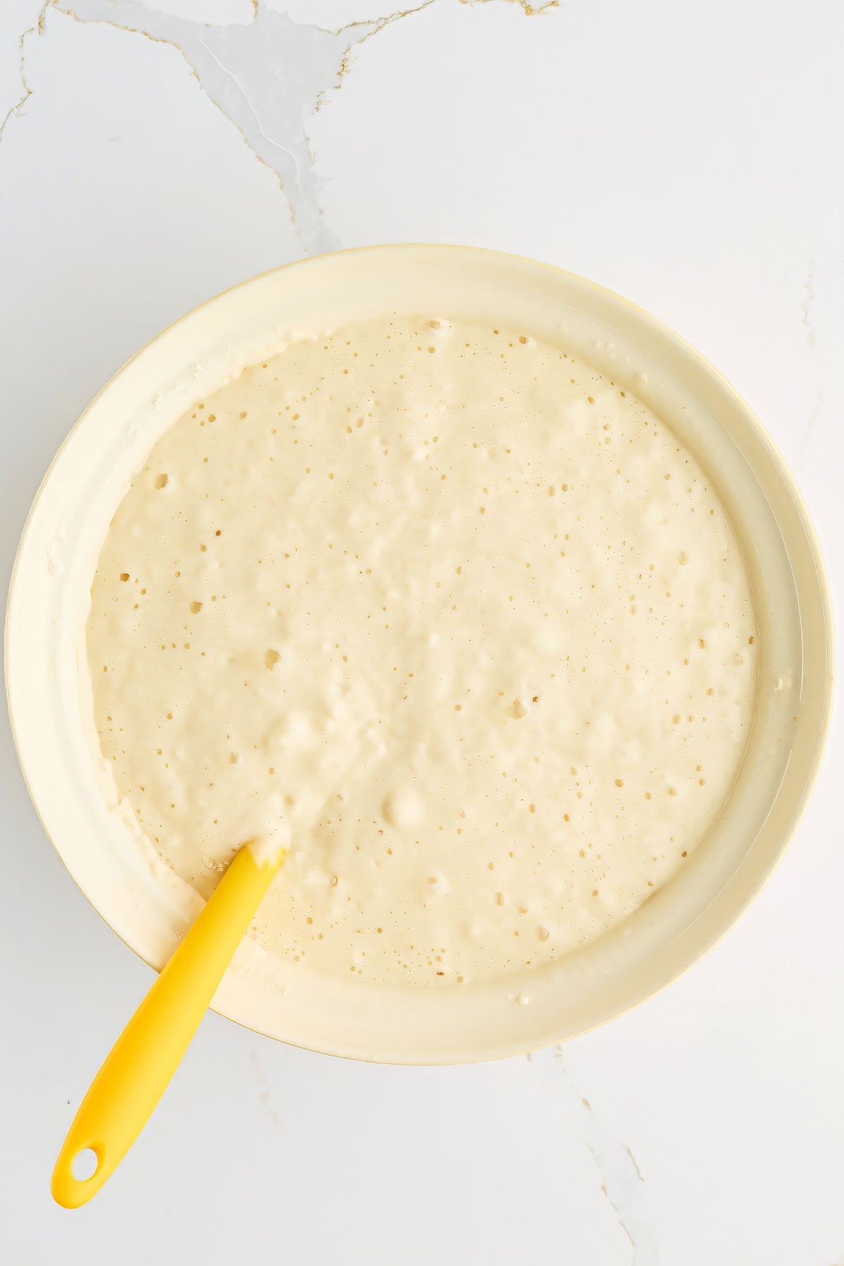 A bowl filled with bubbly pancake batter and a yellow spatula resting inside, placed on a white marble surface.