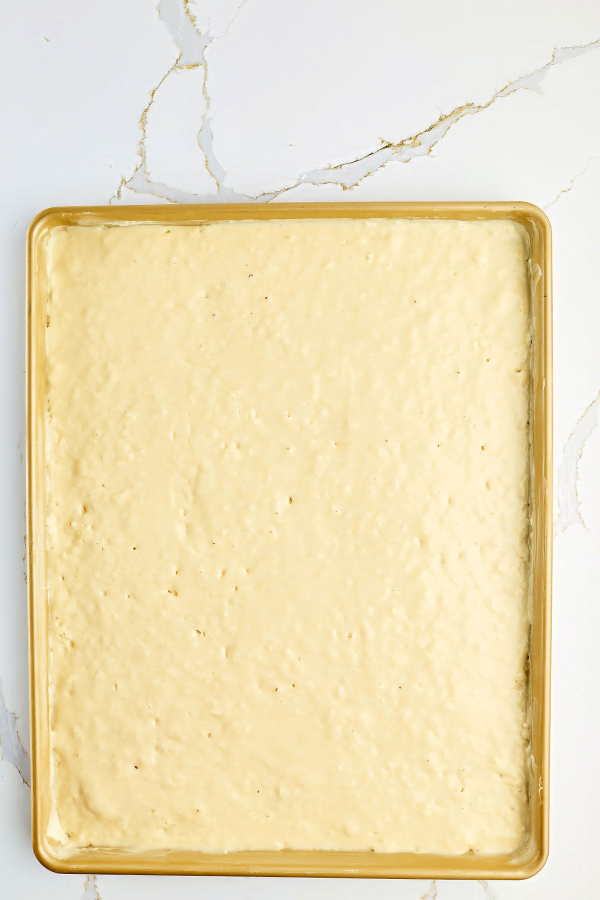 A baking sheet filled with an even layer of unbaked cake batter on a white marble surface.