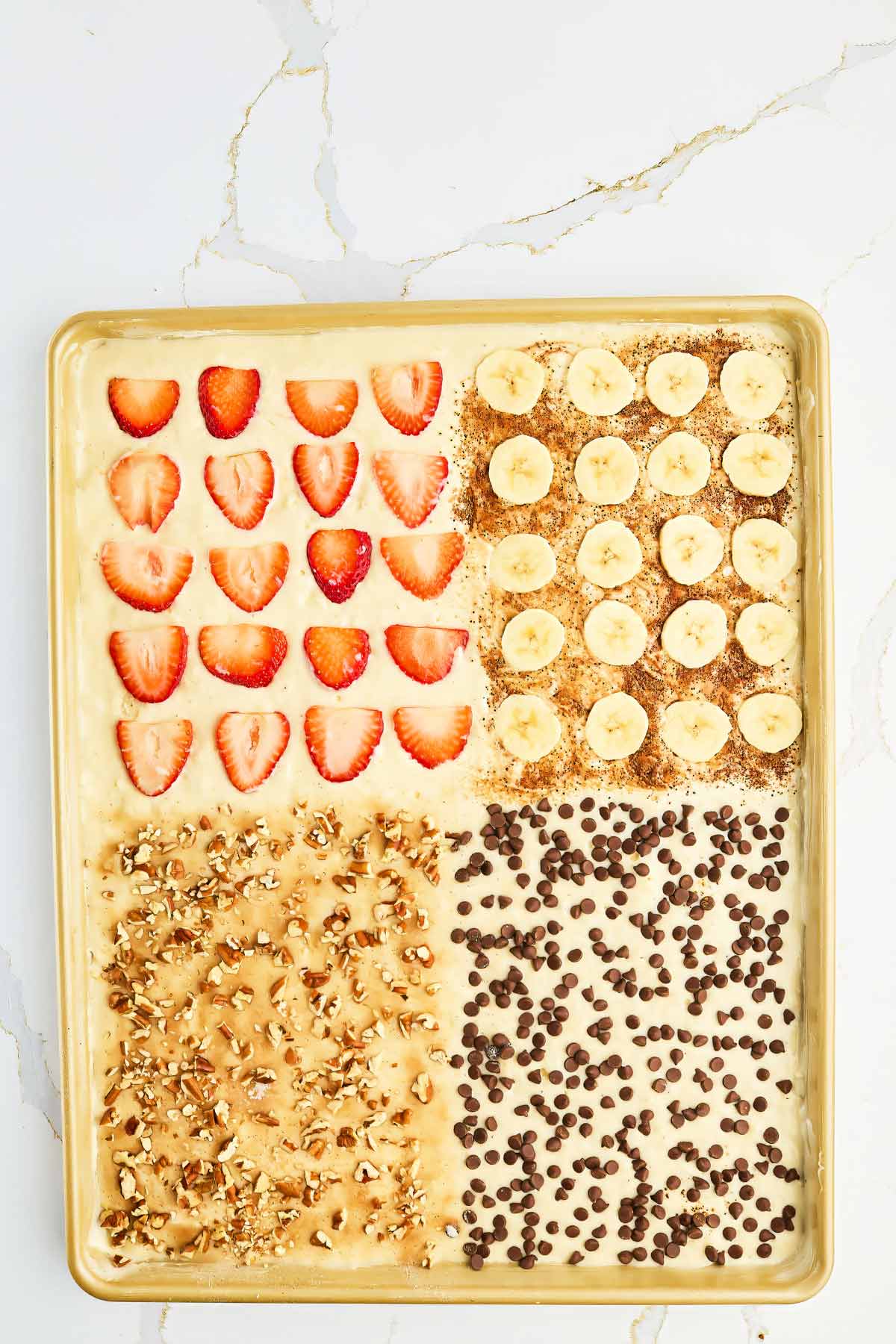 A sheet pan of baked oatmeal topped in four sections with sliced strawberries, sliced bananas with flaxseed, chopped nuts, and chocolate chips on a marble surface.