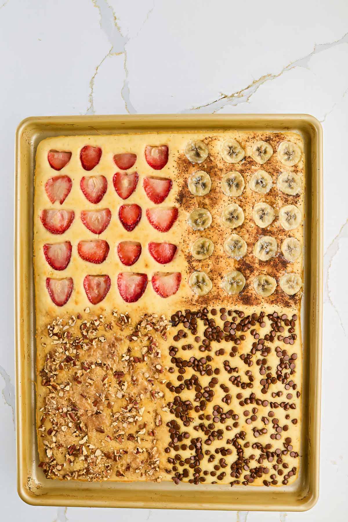 A sheet pan of baked pancake batter divided into four sections, each topped separately with strawberries, banana slices, chopped nuts, and chocolate chips.