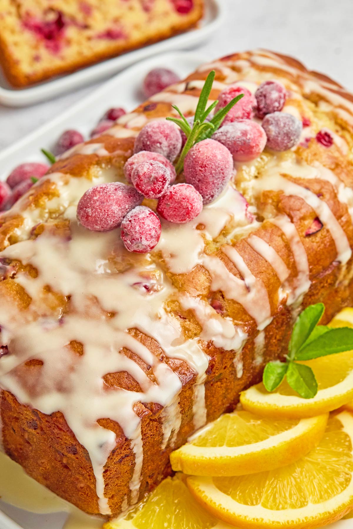 A loaf of glazed cranberry bread garnished with sugared cranberries and rosemary, served with orange slices on the side—perfect alongside easy sheetpan pancakes for a festive breakfast spread.