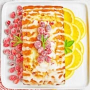 A rectangular loaf cake with white icing is topped with sugared cranberries and rosemary, served on a white plate with sliced lemons and extra cranberries—perfect alongside easy sheetpan pancakes for a festive brunch spread.