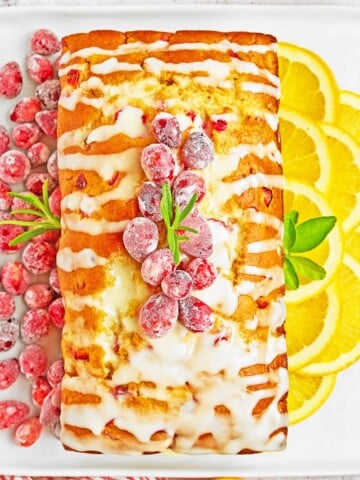 A rectangular loaf cake with white icing is topped with sugared cranberries and rosemary, served on a white plate with sliced lemons and extra cranberries-perfect alongside easy sheetpan pancakes for a festive brunch spread.