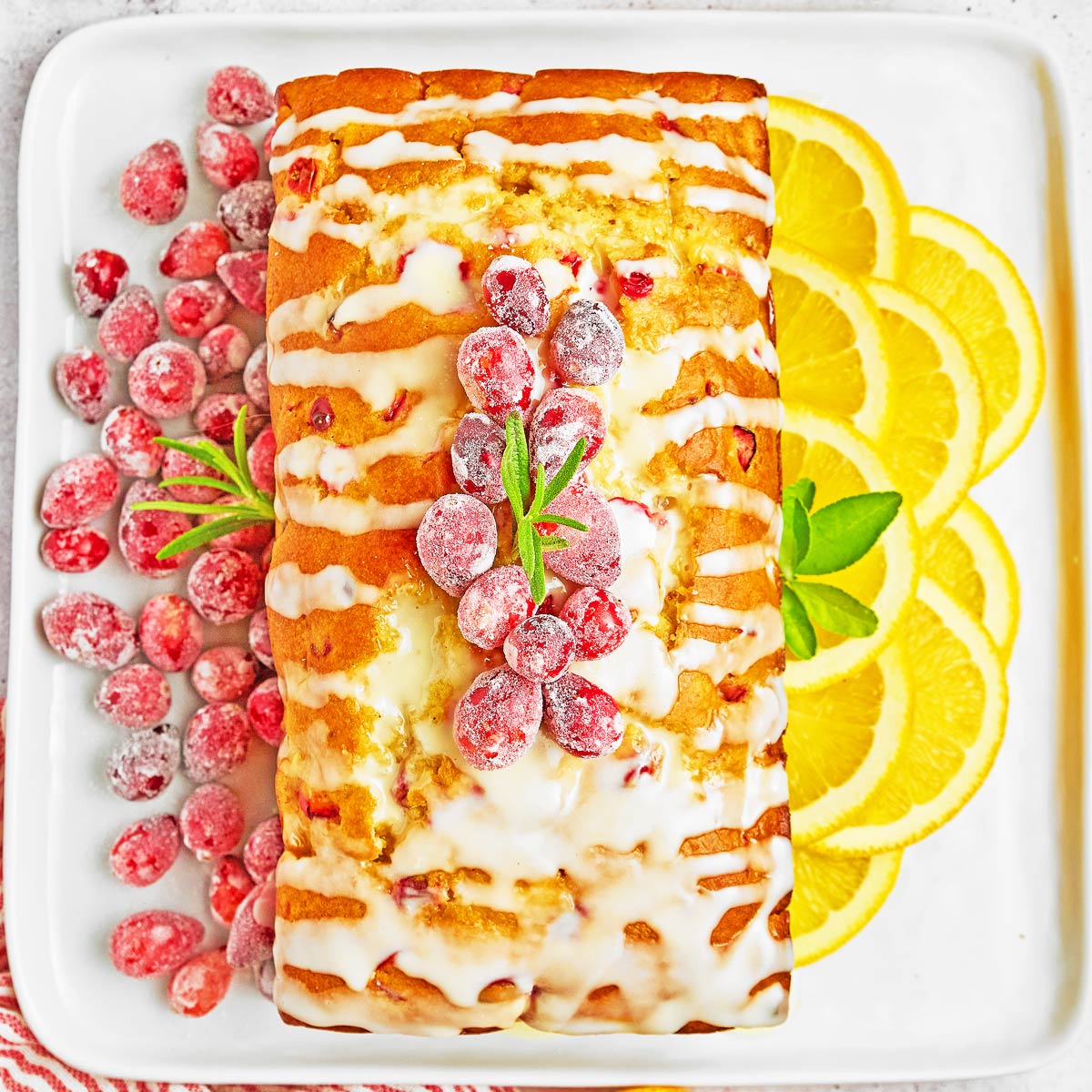 A rectangular loaf cake with white icing is topped with sugared cranberries and rosemary, served on a white plate with sliced lemons and extra cranberries-perfect alongside easy sheetpan pancakes for a festive brunch spread.