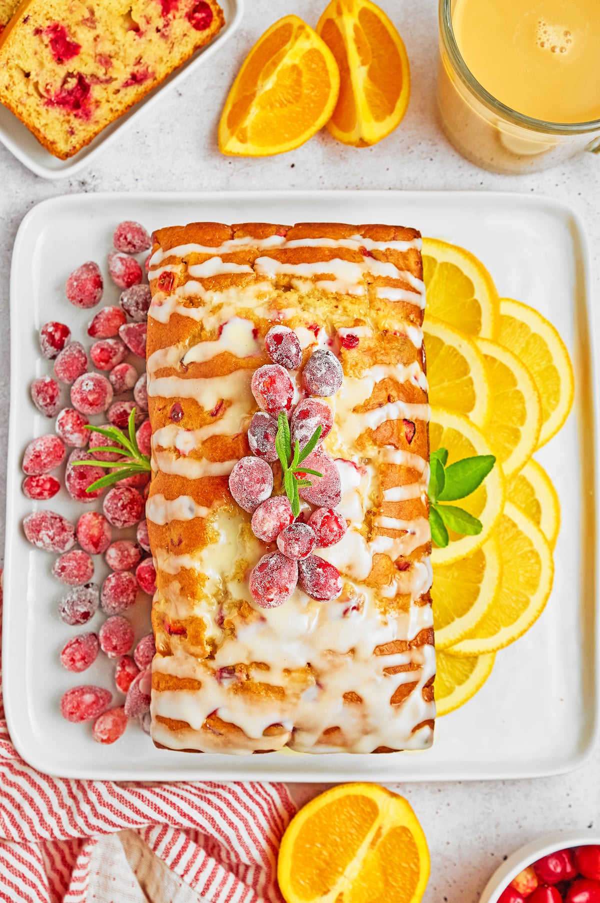 A glazed loaf cake topped with sugared cranberries and mint sits on a white plate, surrounded by orange slices—perfect inspiration for your next brunch alongside sheet pan pancakes from mix.